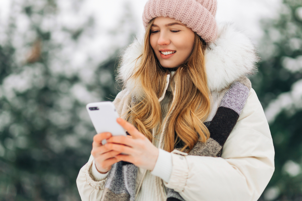 Eine Frau in Winterjacke und Mütze steht in einer Schneelandschaft und nutzt ihr Smartphone.