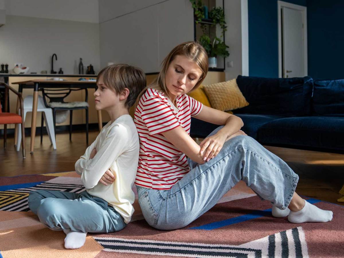Mutter und Sohn sitzen nach einem Streit Rücken an Rücken. Der Sohn schaut starr und verärgert in die Ferne, die Mutter schaut über die Schulter zu ihm.