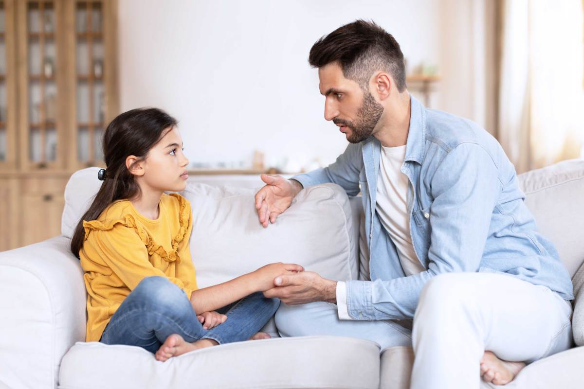 Vater und Tochter sitzen auf dem Sofa. Der Vater hält die Hand seiner Tochter und scheint ihr etwas Ernstes zu erklären.