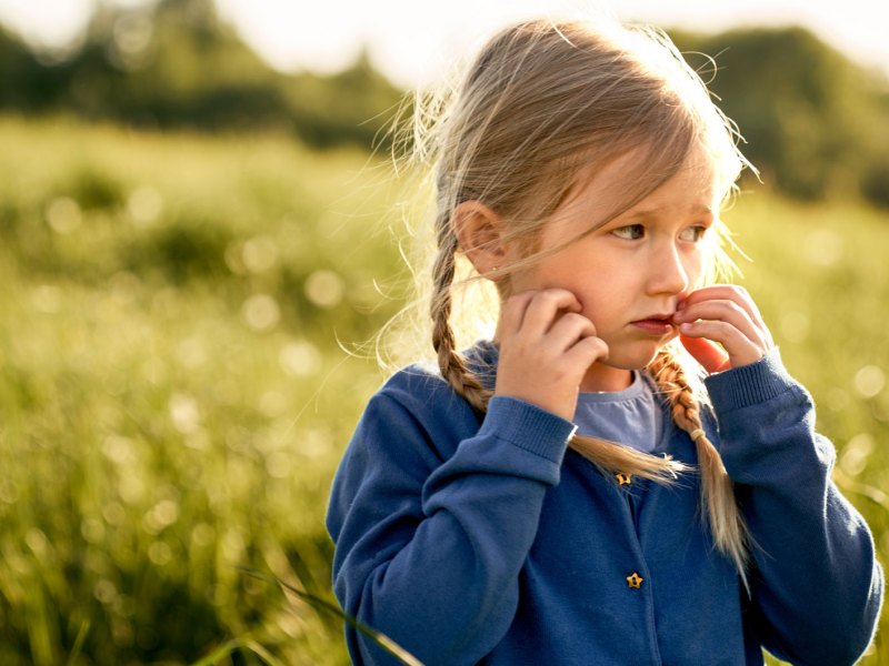 Mädchen, ca. 6 Jahre, steht unsicher auf einer großen Wiese und schaut in die Ferne.