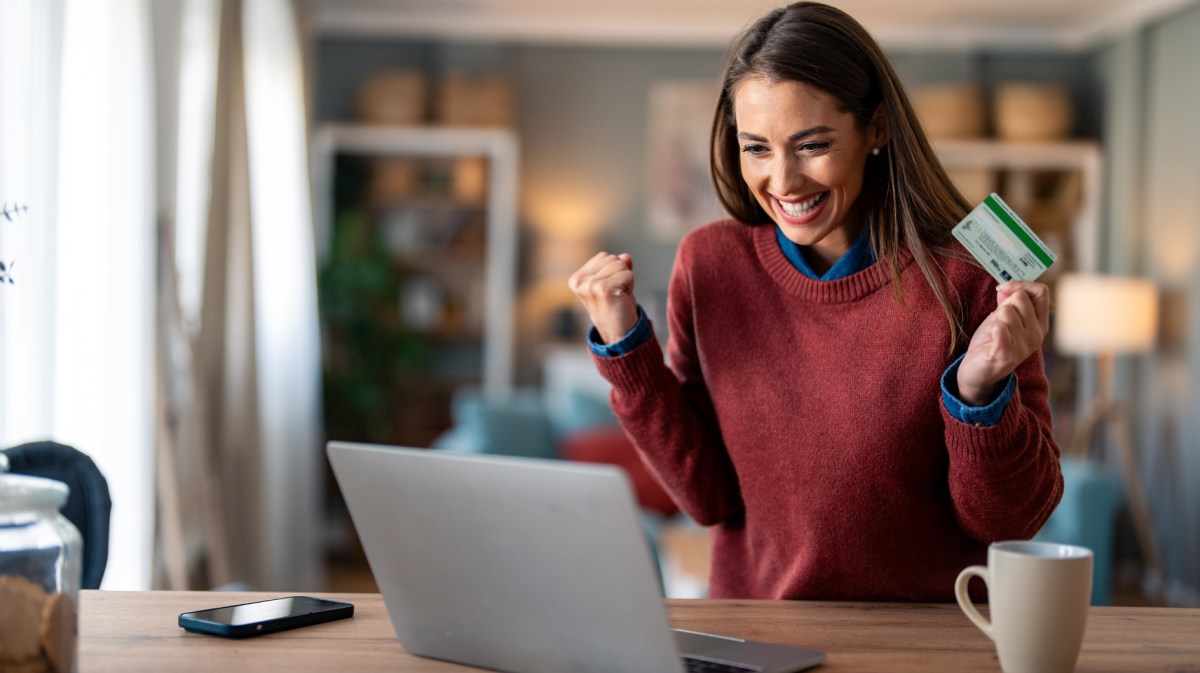 Frau sitzt vor einem Laptop und hält ihre Bankkarte in der Hand. Dabei freut sie sich sehr.