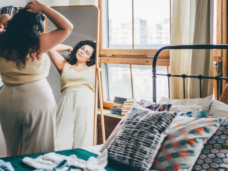 Lockig behaarte übergewichtige junge Frau mit Brille bindet weißes Hemd und bewundert Wahl vor großem Spiegel in stilvollem Zimmer Reflexion Ansicht stehen