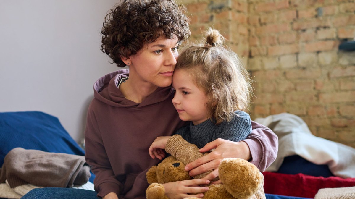 Mutter und Kind sitzen auf einem Bett. Das Lind hält einen Teddy im Arm und kuschelt sich an die Mutter.