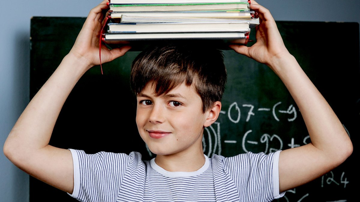 Junge steht vor einer Tafel und hält sich einen Packen Bücher auf dem Kopf