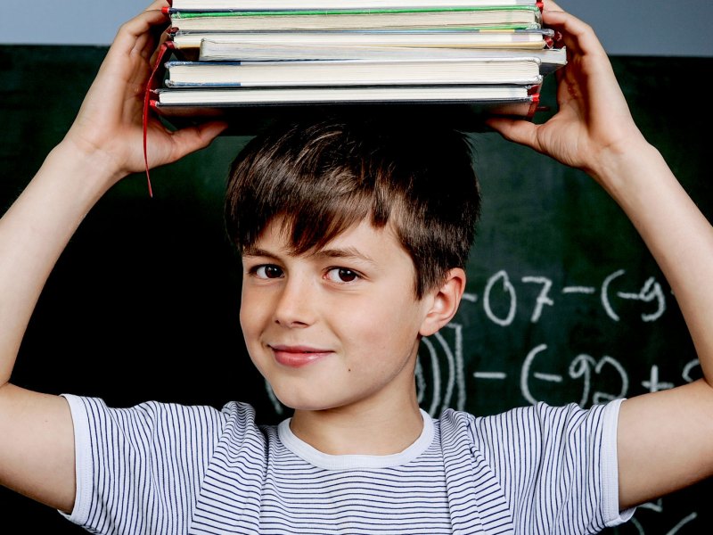 Junge steht vor einer Tafel und hält sich einen Packen Bücher auf dem Kopf