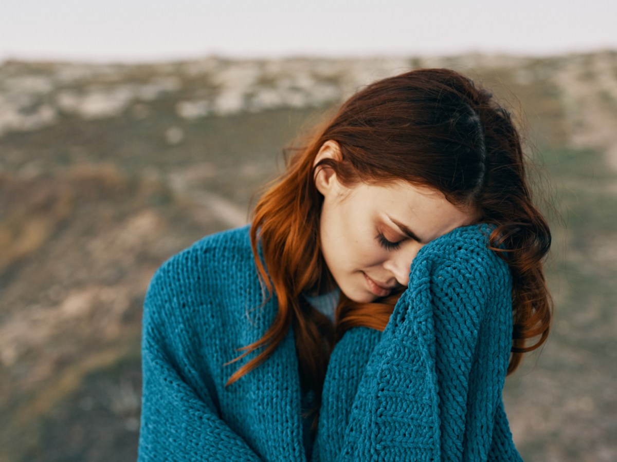 Frau mit blauem Pullover, die draußen steht und sich die Hand vor den Kopf hält