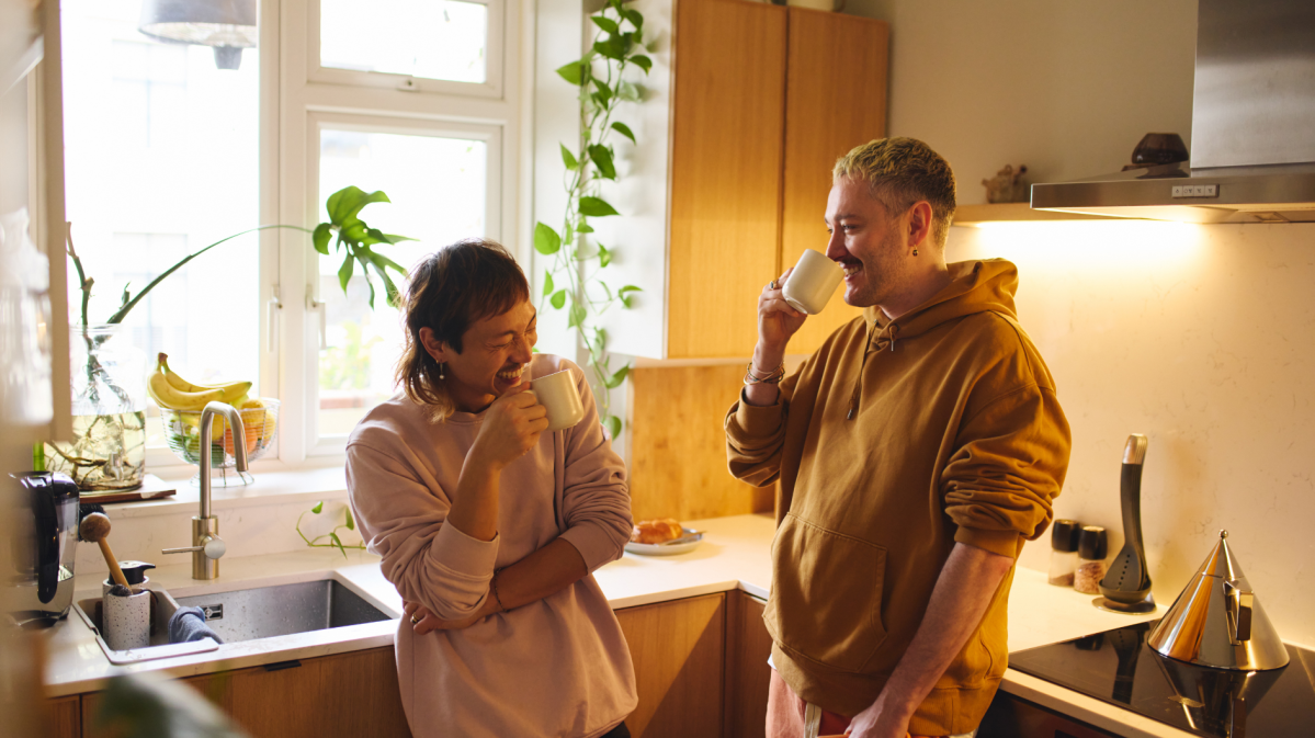 Frau und Mann in der Küche, die beide an einer Tasse nippen und lachen.