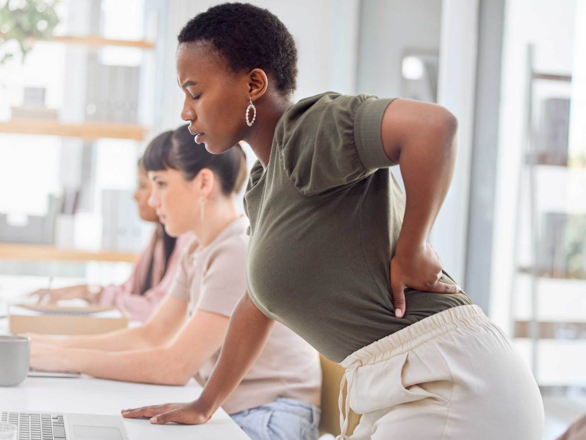 Junge Frau mit Kurzhaarfrisur leidet am Arbeitsplatz sichtlich an Rückenschmerzen.