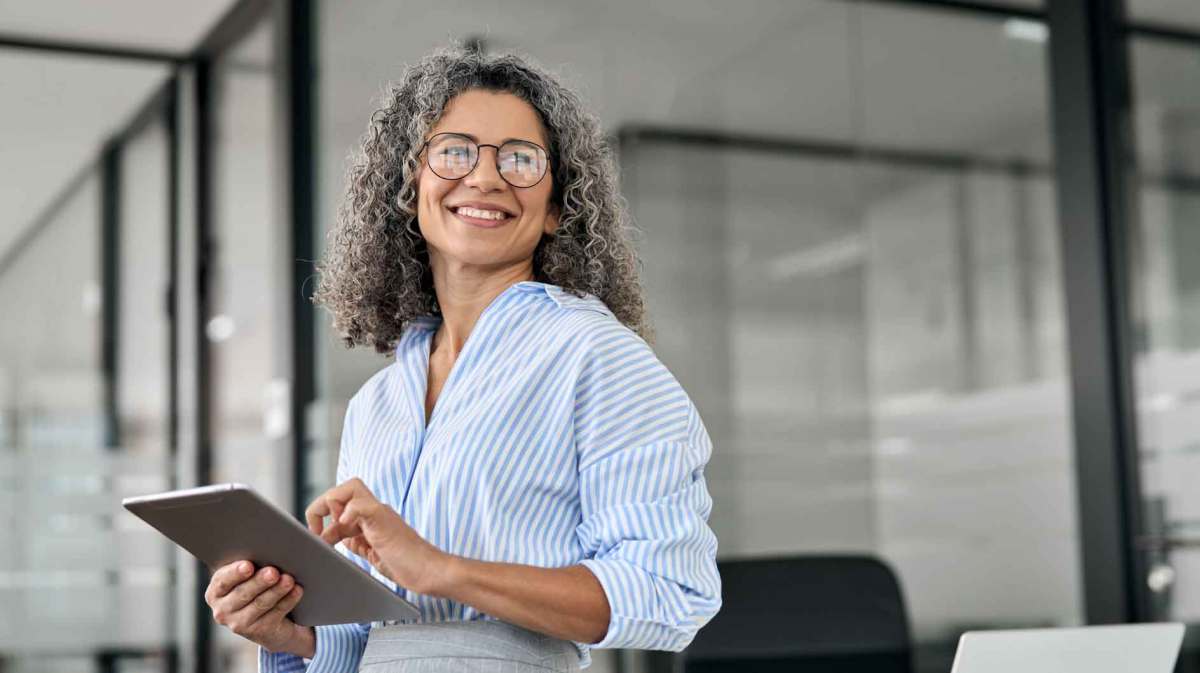 Ü50-Business Women steht auf der Arbeit mit einem Tablet und lächelt.