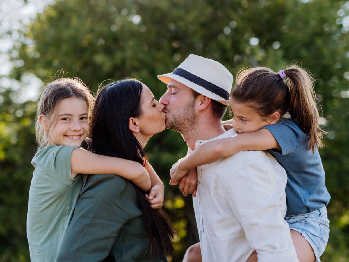 Familie auf einem Spaziergang, Mutter und Vater tragen jeweils eine Tochter Huckepack und geben sich einen Kuss.