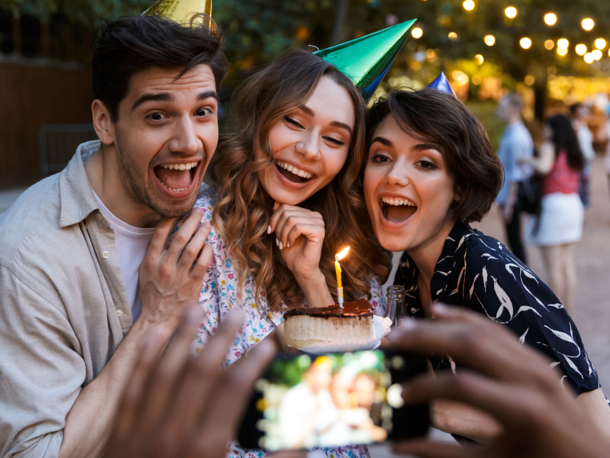 Zwei Frauen und ein Mann mit Partyhut und Kuchen in der Hand, von denen ein Foto gemacht wird