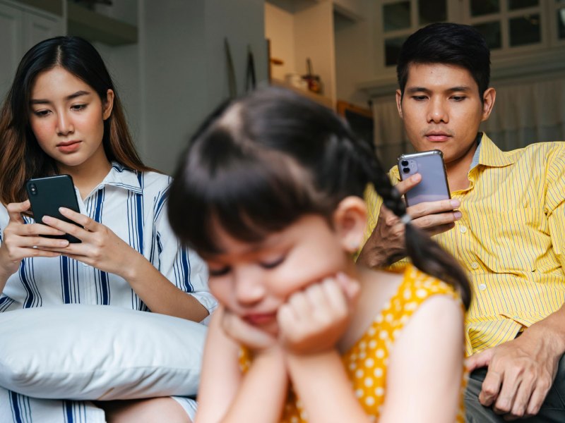 Eltern sitzen mit ihren Smartphones auf dem Sofa, die Tochter hockt gelangweilt im Vordergrund und weiß nichts mit sich anzufangen.