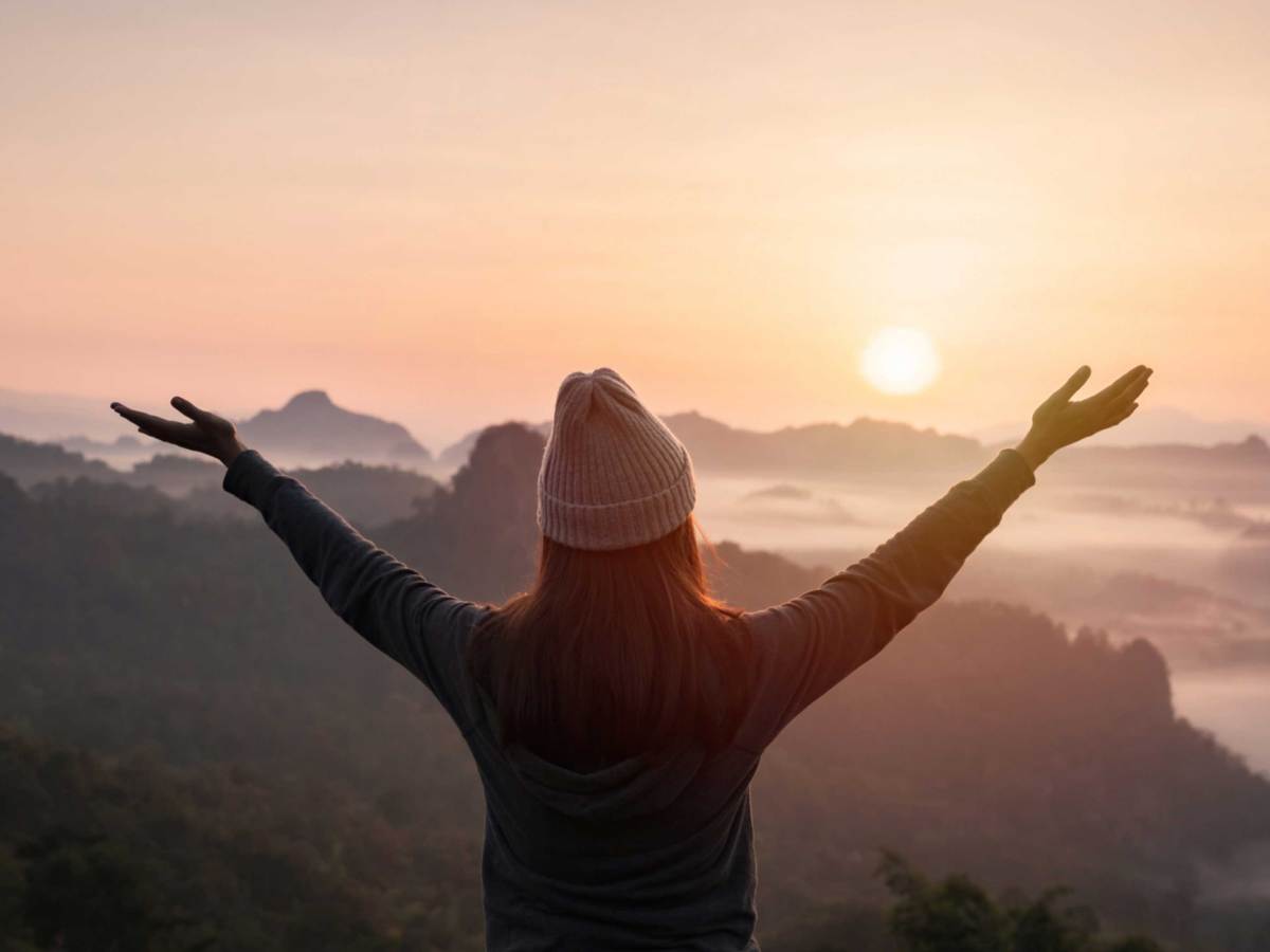 Frau mit M&uuml;tze steht mit ausgestreckten Armen vor einer Berglandschaft