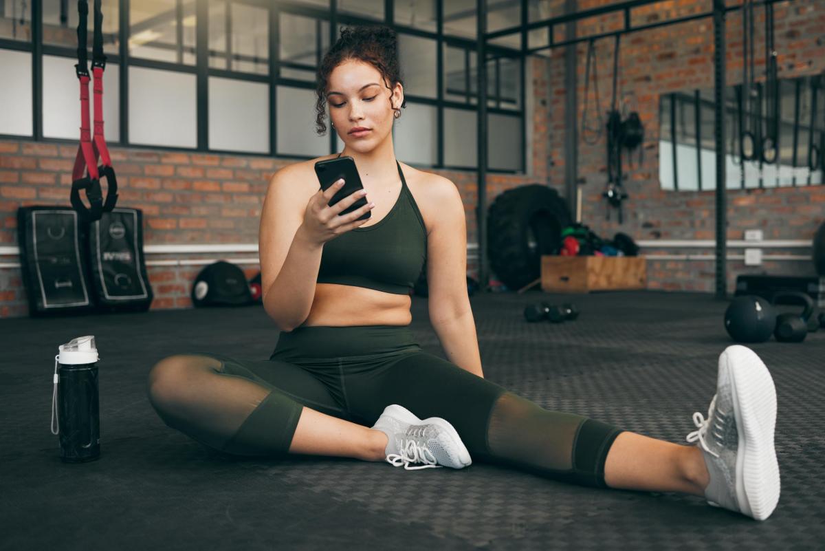 Frau im Fitnessstudio beim Workout, mit Smartphone und Fitness App. Junge Sportlerin mit Smartphone für Fortschritt, Leistung und Kommunikation für einen gesunden Lebensstil