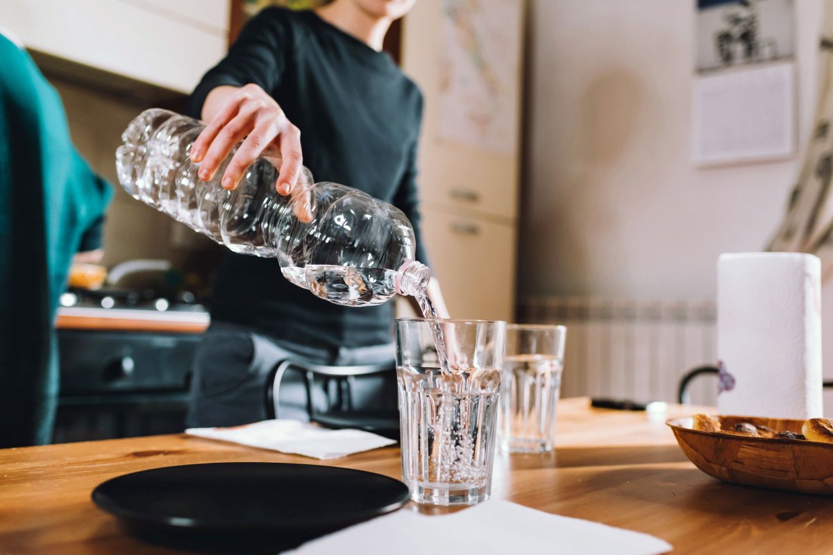 Frau schüttet Wasser aus Wasserflasche in ein Glas, dahinter ist eine Küche