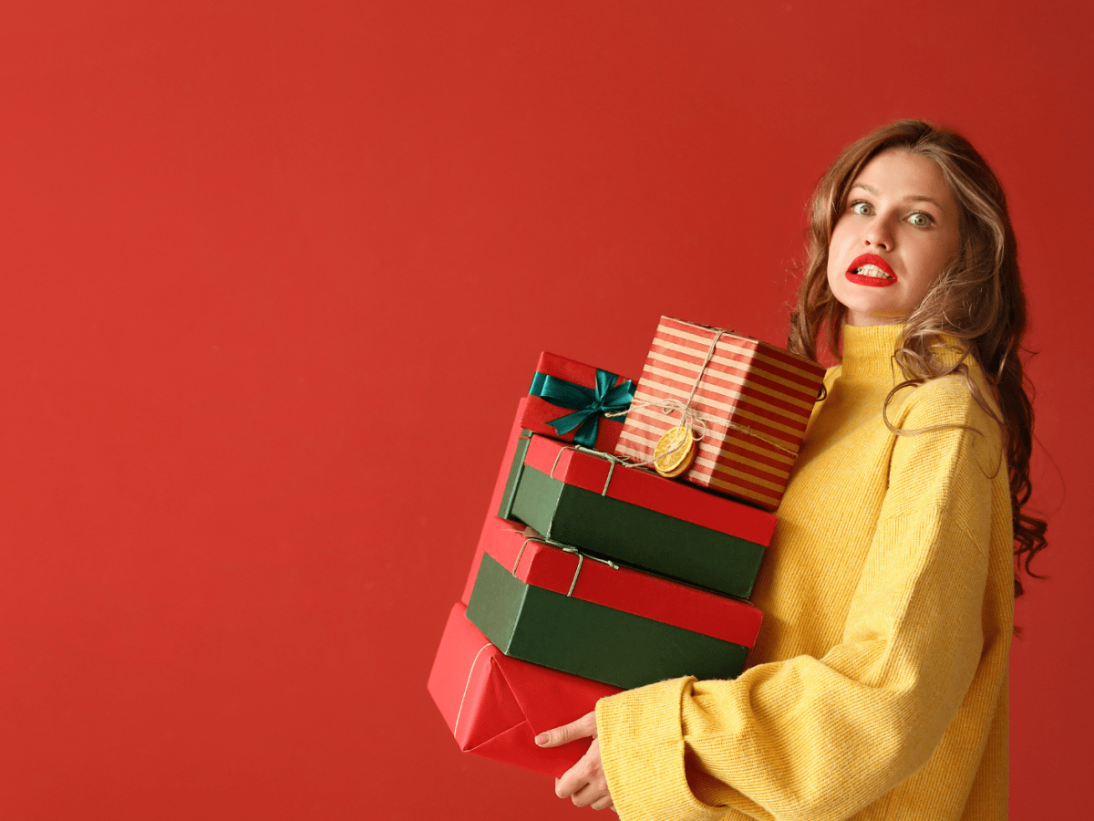 Junge Frau mit langen braunen Haaren und rotem Lippenstift trägt stapelweise bunt verpackte Geschenke vor rotem Hintergrund.