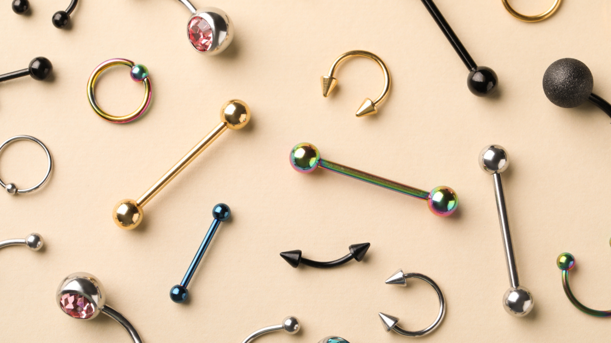 Verschiedene Piercingschmuckstücke in Gold, Silber, Schwarz und Regenbogenfarben liegen dekorativ auf einem beigen Hintergrund.