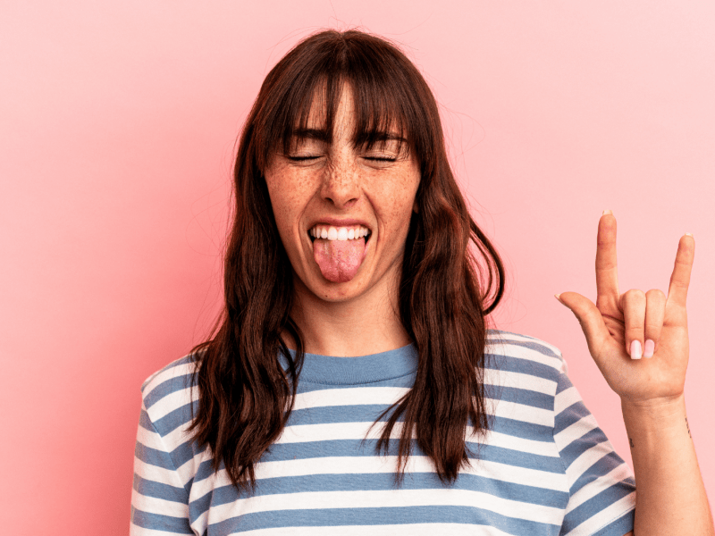 Junge Frau mit Sommersprossen und braunem Haar streckt die Zunge heraus und zeigt die Mano-Cornuta Geste vor rosa Hintergrund.