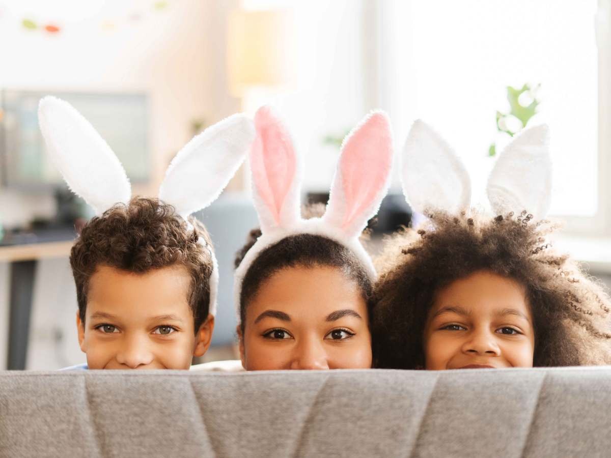 Mutter und zwei Kinder schauen mit Hasenohren auf dem Kopf verschmitzt über die Sofalehne.