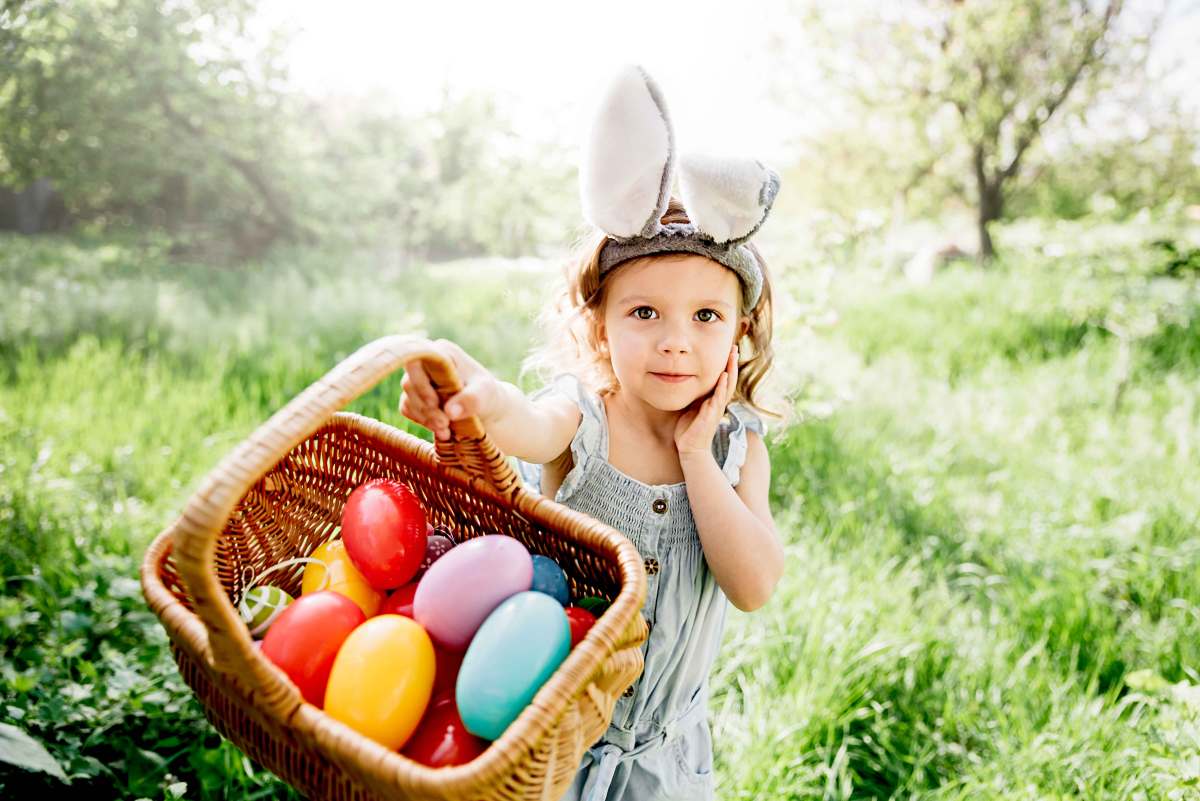 Kleines Mädchen mit Hasenohren auf dem Kopf zeigt stolz ihren prall gefüllten Osterkorb in die Kamera.