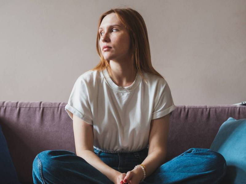 Frau sitzt im Schneidersitz auf einer Couch und guckt betr&uuml;bt zur Seite.
