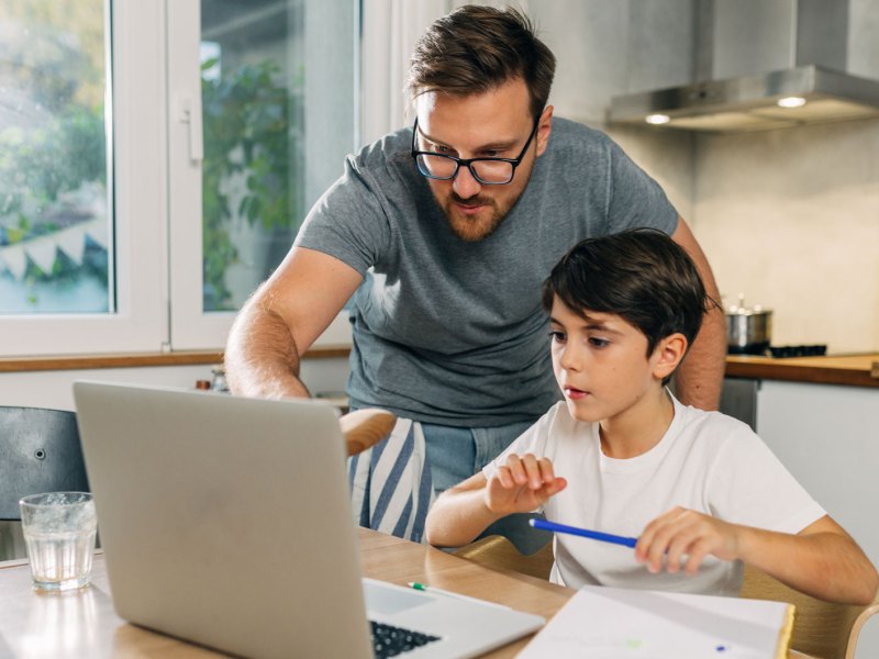 Vater und Sohn in der K&uuml;che am Laptop. Der Vater hilft seinem Sohn bei den Hausaufgaben.