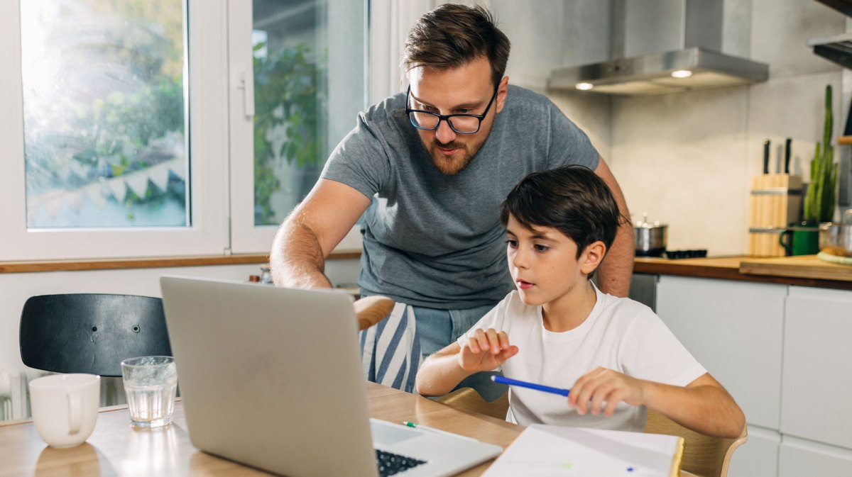 Vater und Sohn in der Küche am Laptop. Der Vater hilft seinem Sohn bei den Hausaufgaben.