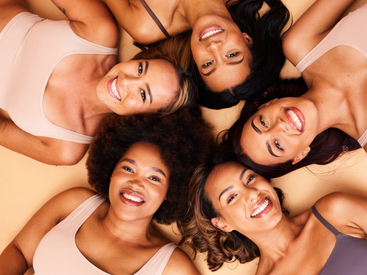 5 Frauen in einem Kreis, die auf dem Boden liegen, ihre K&ouml;pfe aneinander halten und in die Kamera l&auml;cheln.