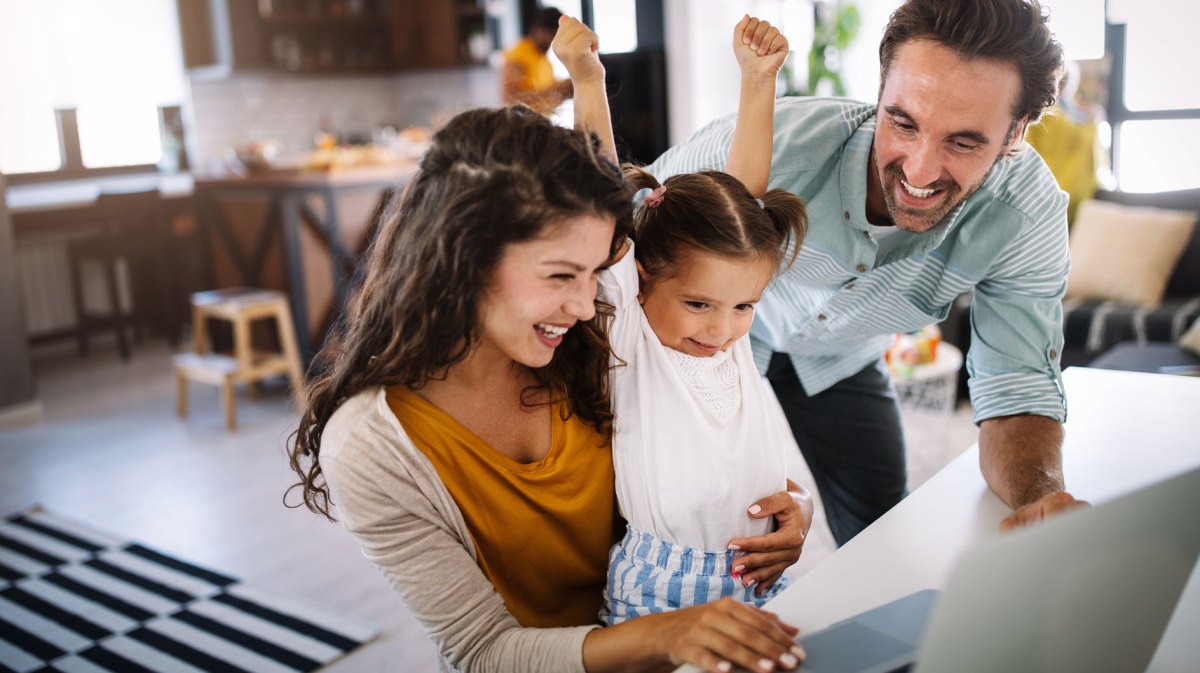 Dreiköpfige Familie aus Mutter, Vater und Tochter sitzen gemeinsam am PC und freuen sich über etwas, was sie auf dem Bildschirm sehen.