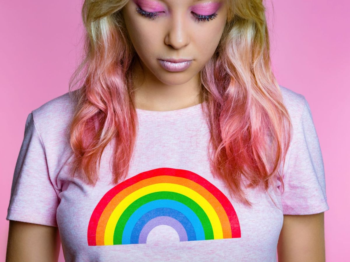 Eine junge Frau mit pinken Haaren vor rosa Hintergrund trägt ein rosa Shirt mit einem Regenbogen