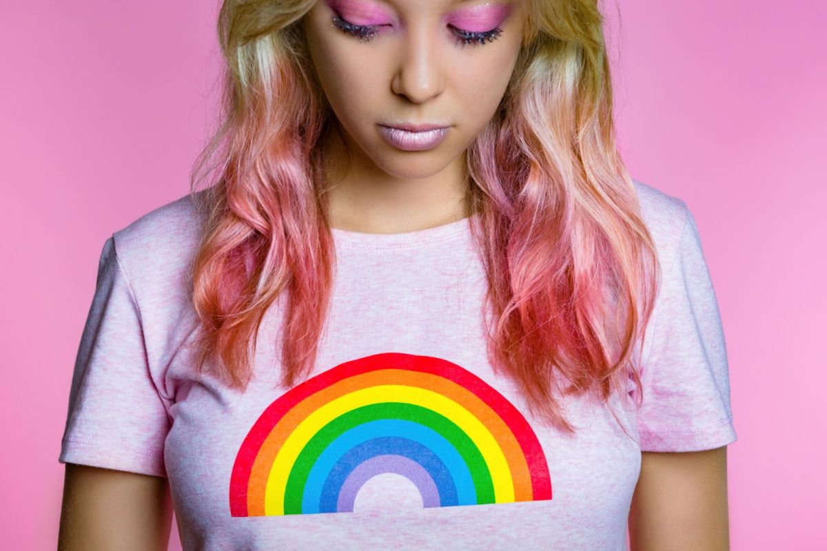 Eine junge Frau mit pinken Haaren vor rosa Hintergrund trägt ein rosa Shirt mit einem Regenbogen