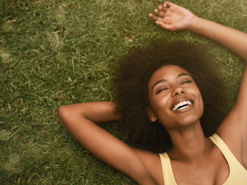 Frau auf einer grünen Wiese, die lächelnd in den Himmel blickt und auf ihren Händen liegt.