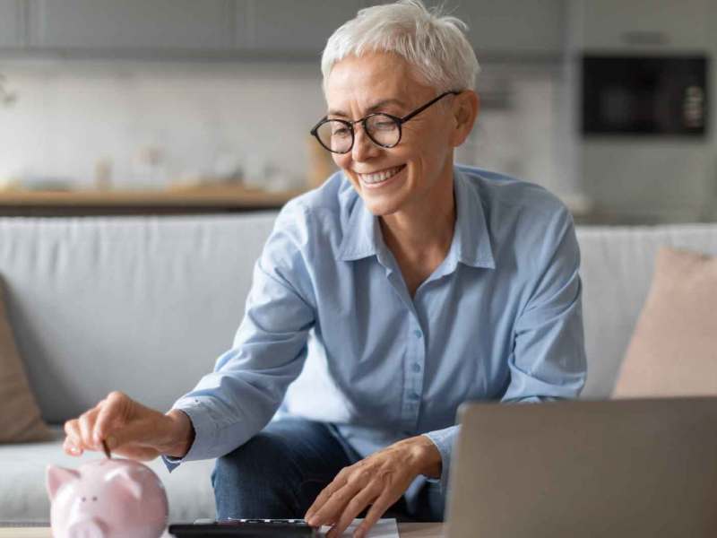 Frau mittleren Alters mit weißen kurzen Haaren sitzt vor einem Notebook und wirft Geld in ein Sparschwein.