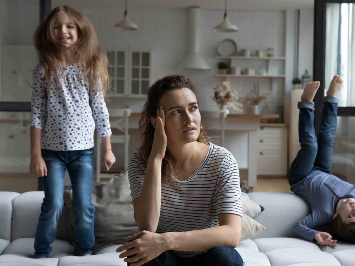 Mutter sitzt genervt auf dem Sofa, w&auml;hrend ihre beiden Kinder um sie herum turnen.