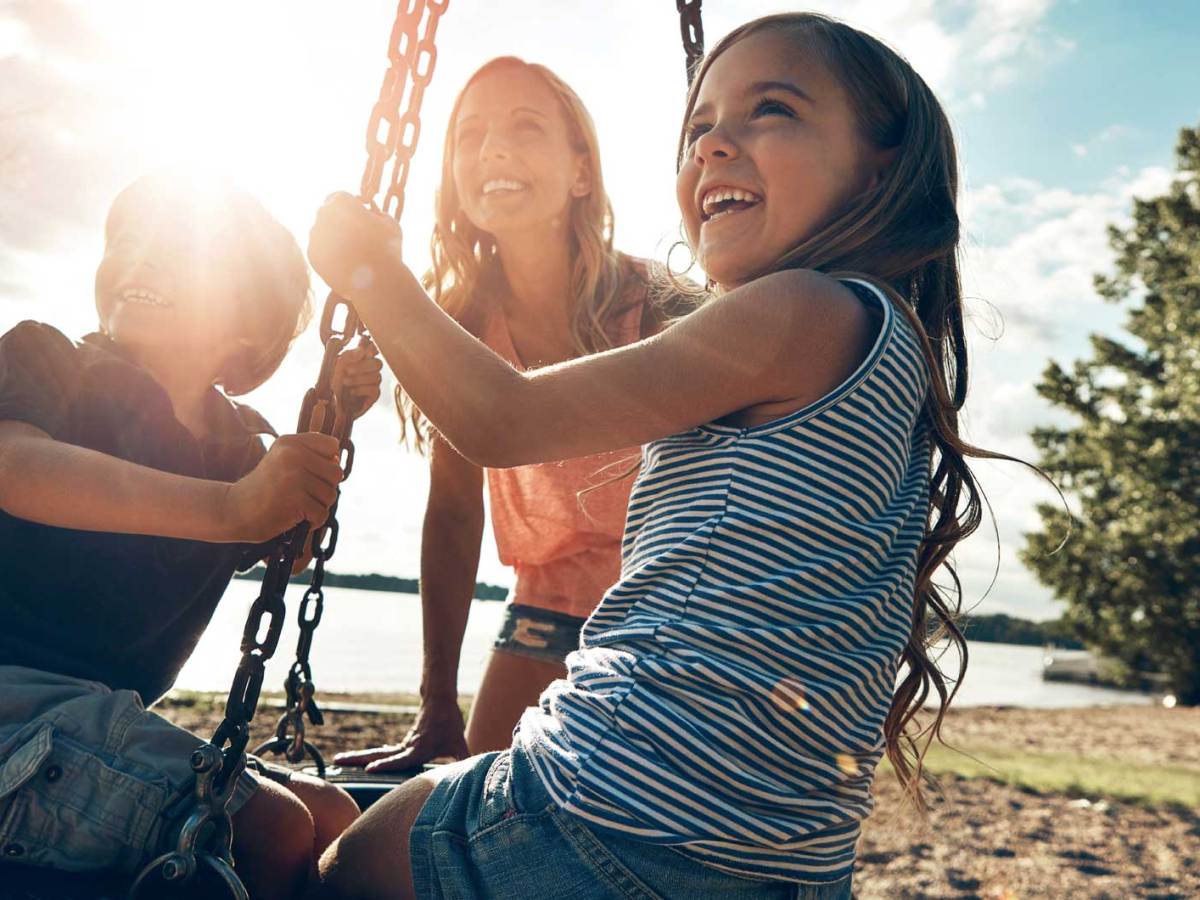 Mutter spielt mit Tochter und Sohn auf einem Spielplatz. Die Kinder sitzen auf einer Reifenschaukel.