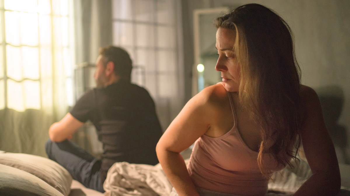 Eine nachdenkliche Frau in einem rosa Top sitzt auf einem Bett, während ein Mann in Schwarz mit dem Rücken zu ihr abgewandt auf die helle Fensterfront blickt.