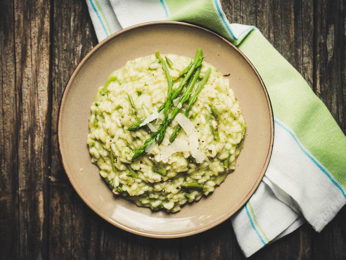 Cremiges Spargelrisotto mit geriebenem Parmesan auf einem braunen Teller, angerichtet auf einem dunklen Holztisch mit gr&uuml;n-wei&szlig;er Serviette.