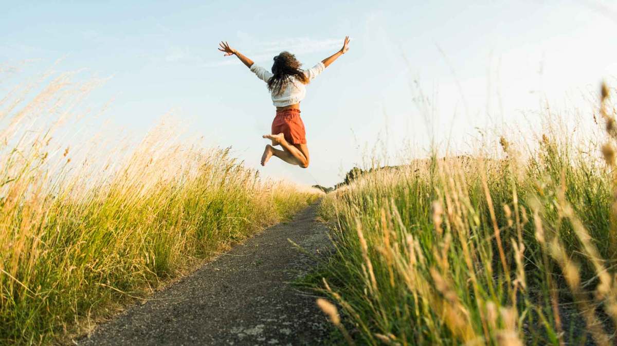 Frau springt auf einem Weg zwischen Wiesen und Feldern nach oben.