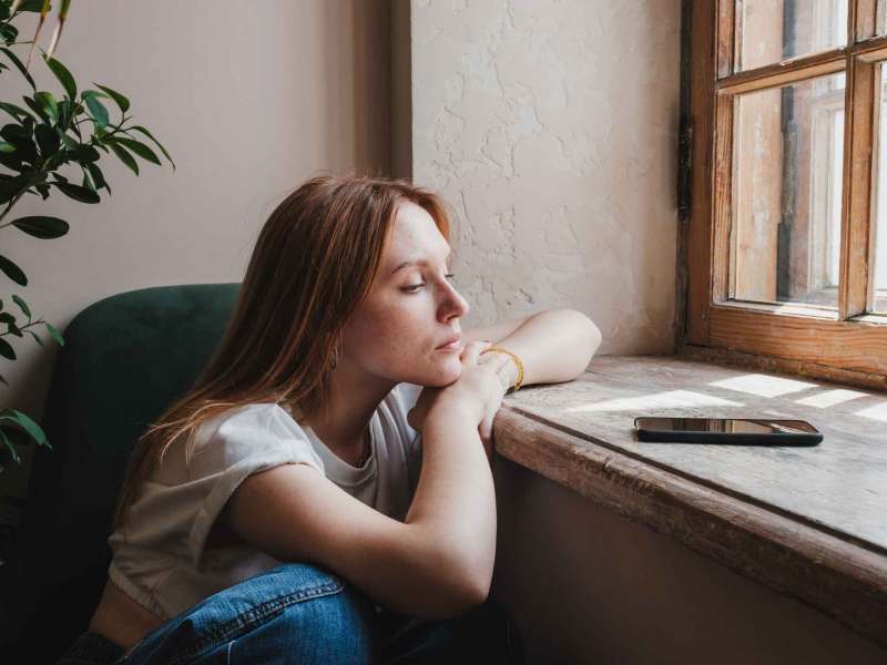 Frau sitzt traurig an der Fensterbank und blickt nach draußen.