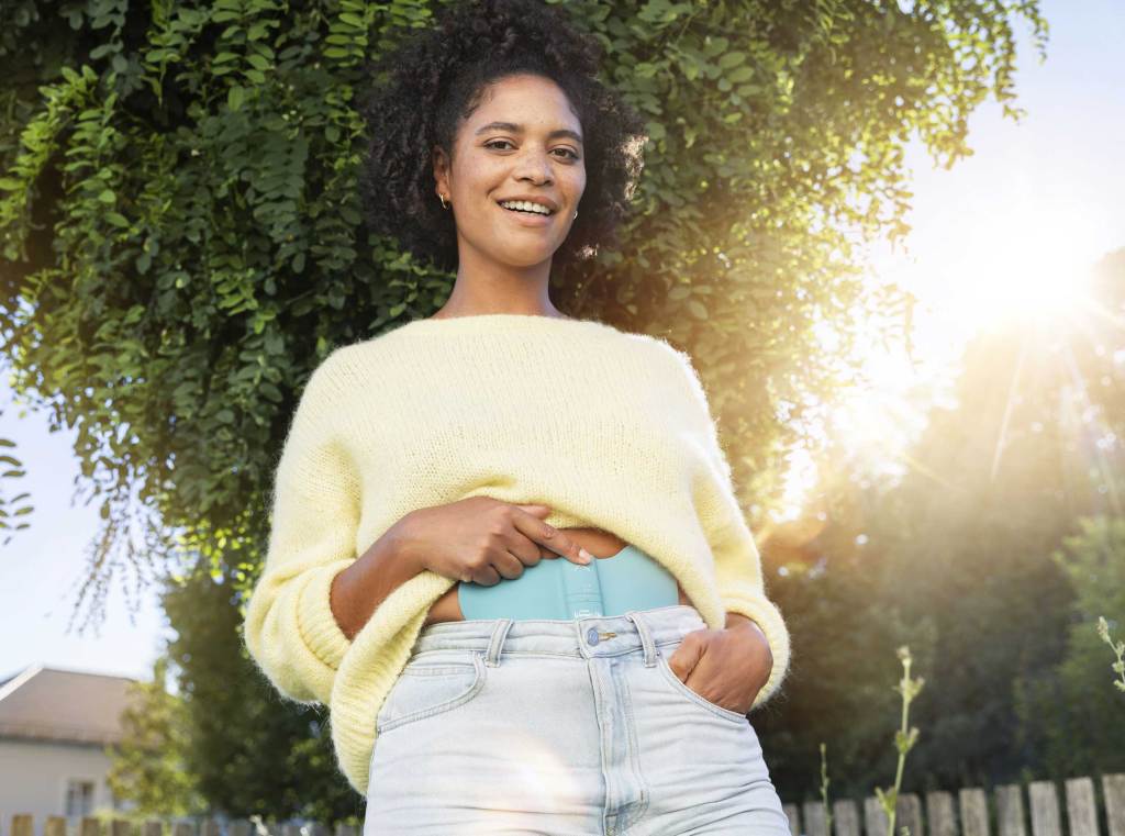 Lächelnde Frau mit lockigem Haar, in gelbem Pullover und heller Jeans steht bei Sonnenschein im Grünen und zeigt ein blaues Wärmepflaster auf dem Bauch.