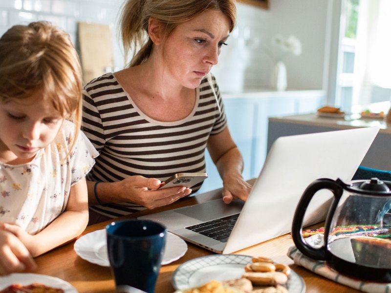 Frau sitzt während des Frühstücks mit der Familie am Laptop.- Vor ihr auf dem Tisch steht eine große Kanne Kaffee.