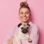 Frau vor einer pinken Wand, mit einem Mops in der Hand, die in die Kamera l&auml;chelt.