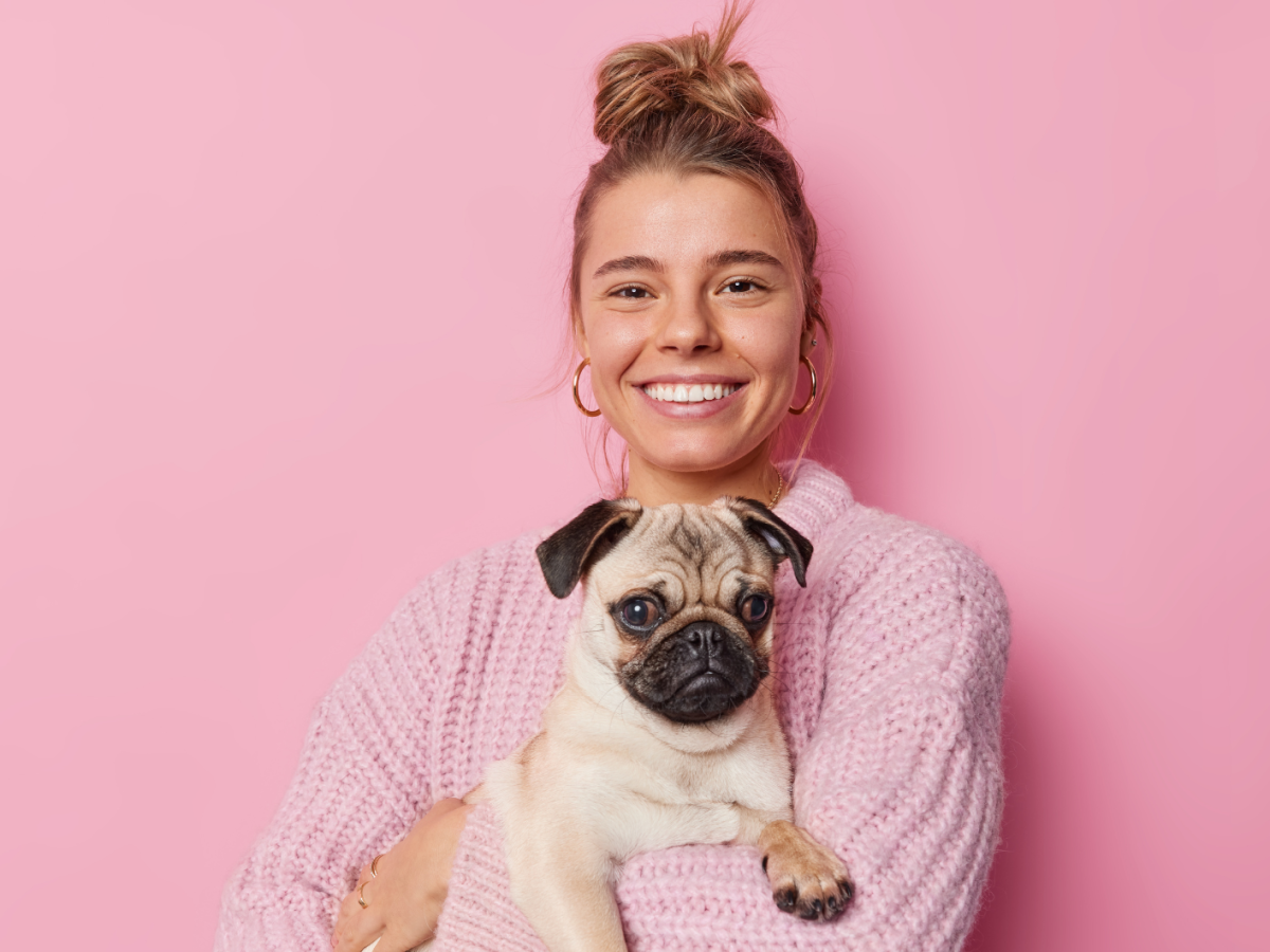 Frau vor einer pinken Wand, mit einem Mops in der Hand, die in die Kamera l&auml;chelt.
