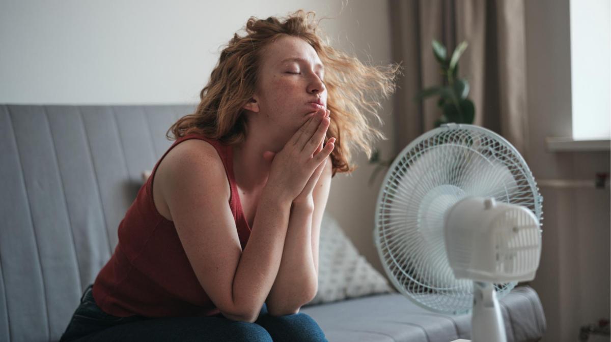 Frau sitzt auf der Couch und kühlt ihr Gesicht mit einem Ventilator.