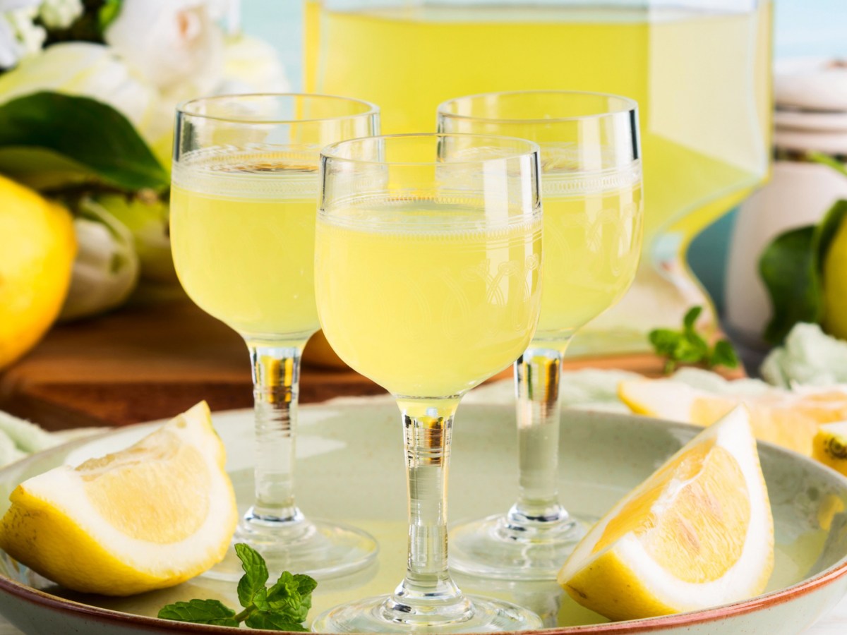 Drei kleine Gl&auml;ser mit Limoncello stehen vor einer gro&szlig;en Flasche, umgeben von Zitronenspalten.