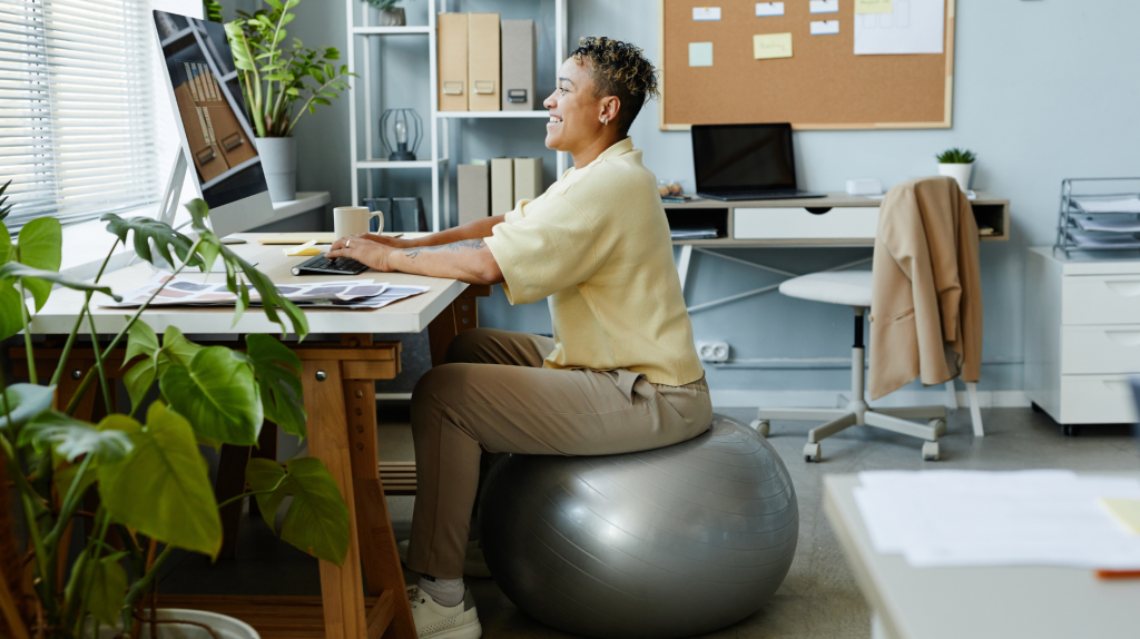 Seitenansicht Porträt der schwarzen jungen Frau sitzt auf Fitness-Ball am Arbeitsplatz und lächelnd, sportliche Aktivität im Büro