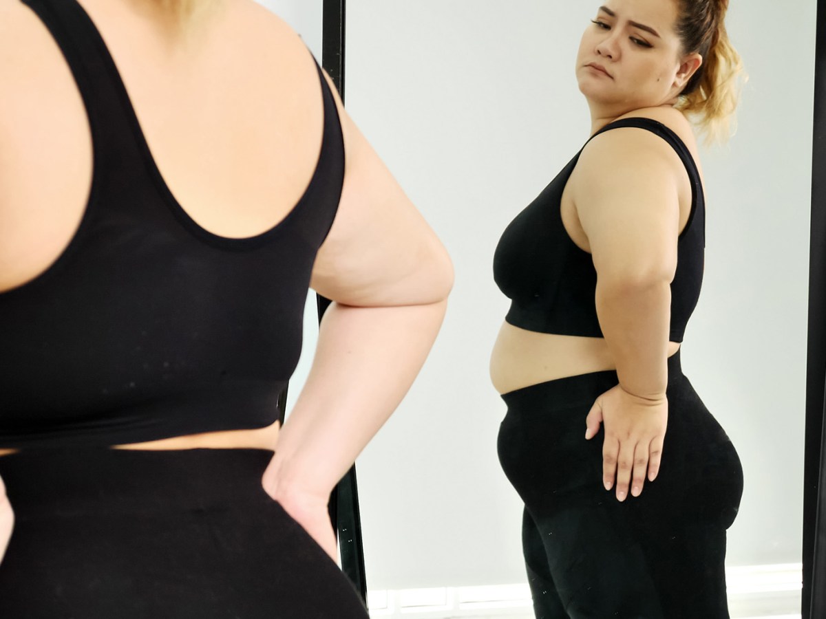 Eine junge Plus-Size-Frau in schwarzem Sport-BH und Leggings schaut kritisch auf ihren Körper im Spiegel.