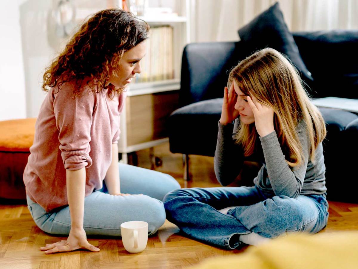 Mutter und Tochter sitzen im Wohnzimmer auf dem Fußboden und unterhalten sich. Die Tochter sieht genervt aus.