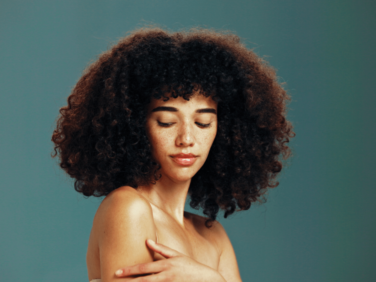 Junge Frau mit lockigem, dunklem Haar und Sommersprossen blickt mit geschlossenen Augen nachdenklich, vor schlichtem, gr&uuml;nlichem Hintergrund.