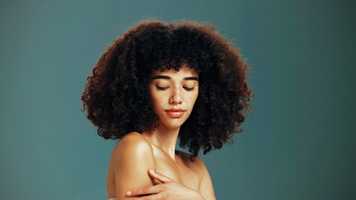 Junge Frau mit lockigem, dunklem Haar und Sommersprossen blickt mit geschlossenen Augen nachdenklich, vor schlichtem, grünlichem Hintergrund.